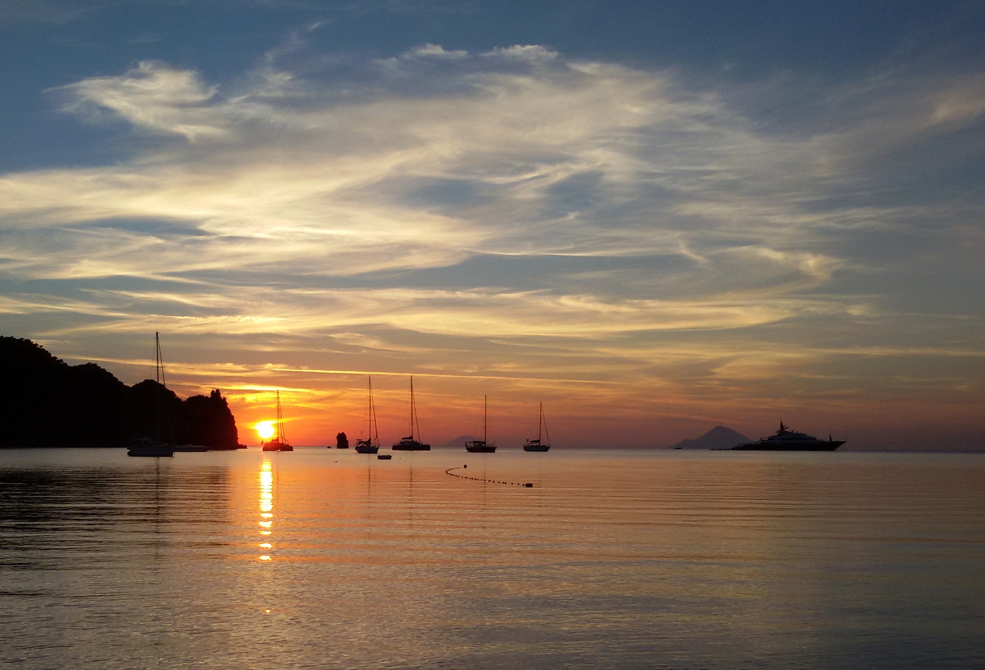 Zu sehen ist der tiefrote Sonnenuntergang über den Liparischen Inseln am Horizont und den Booten, die auf dem ruhigen Meer ankern.